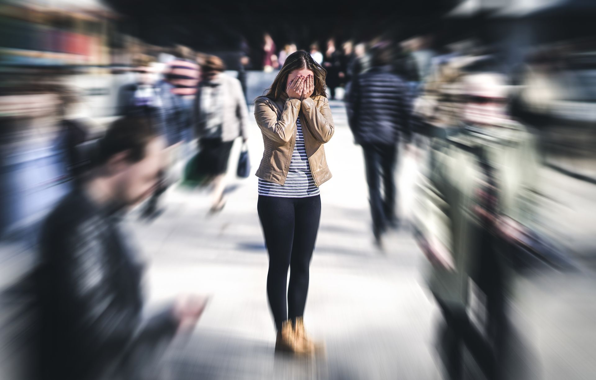 Blurred crowd surrounds a centered person standing on a city street, hands covering their face.