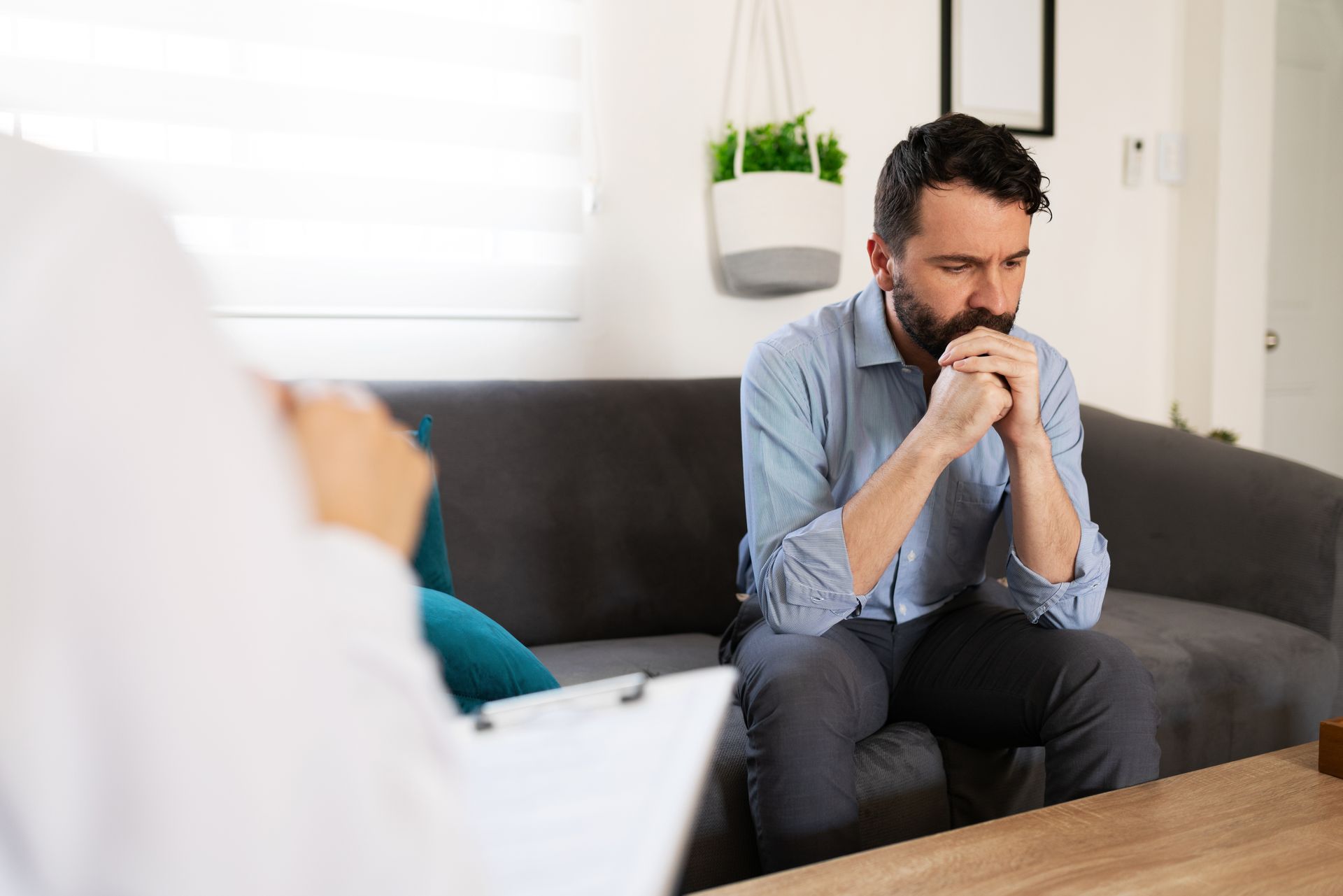 Man sitting on a couch, hands clasped near his face, in a bright 상담 room with a therapist nearby