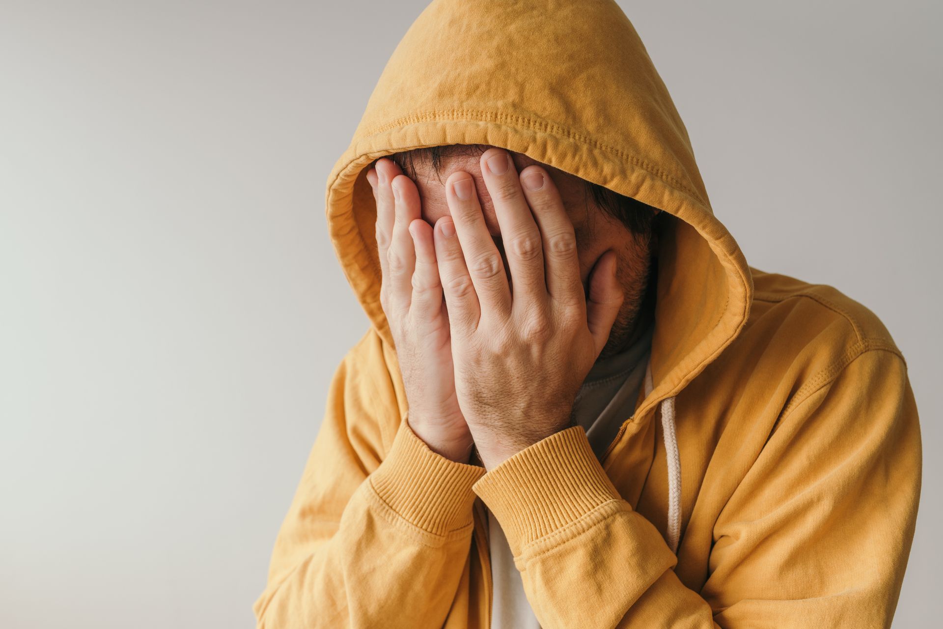 Person in a yellow hoodie covering their face with both hands against a plain background