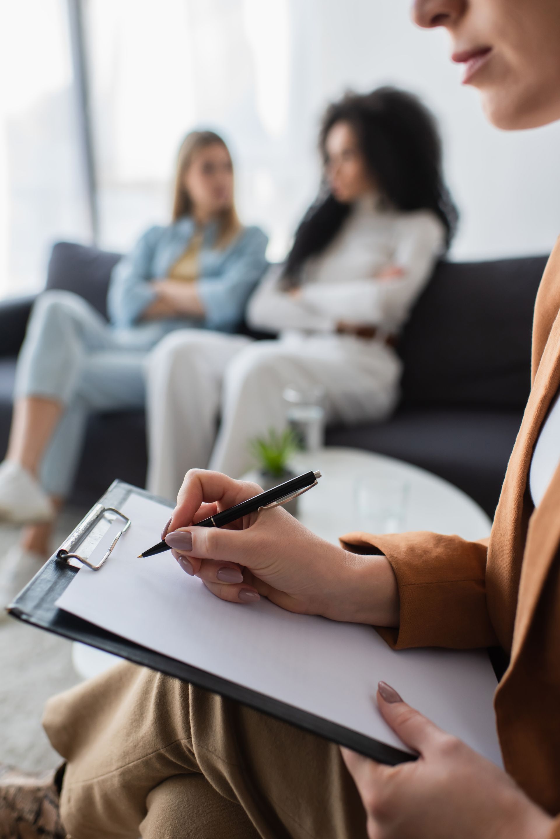 Counselor taking notes on a clipboard during a therapy session with two seated clients in the background