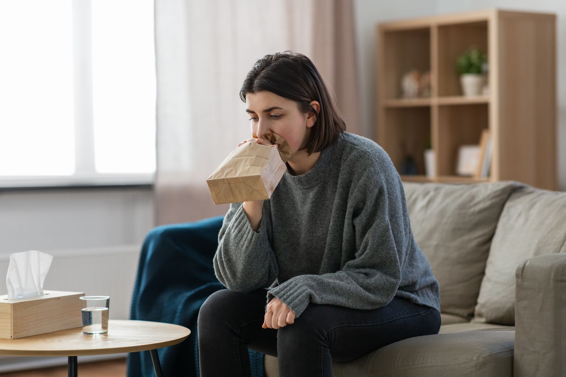 Woman seated on a couch, smelling a tissue box in a bright living room.