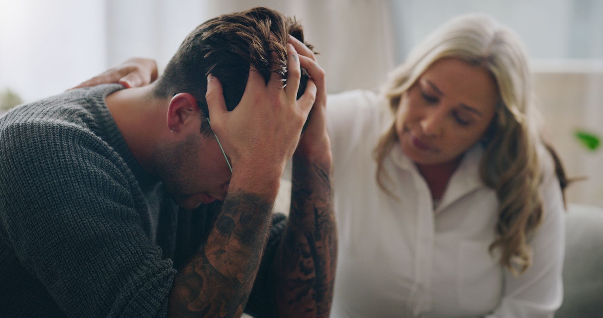 Distressed man sits with head in hands while a concerned woman leans toward him on a couch.