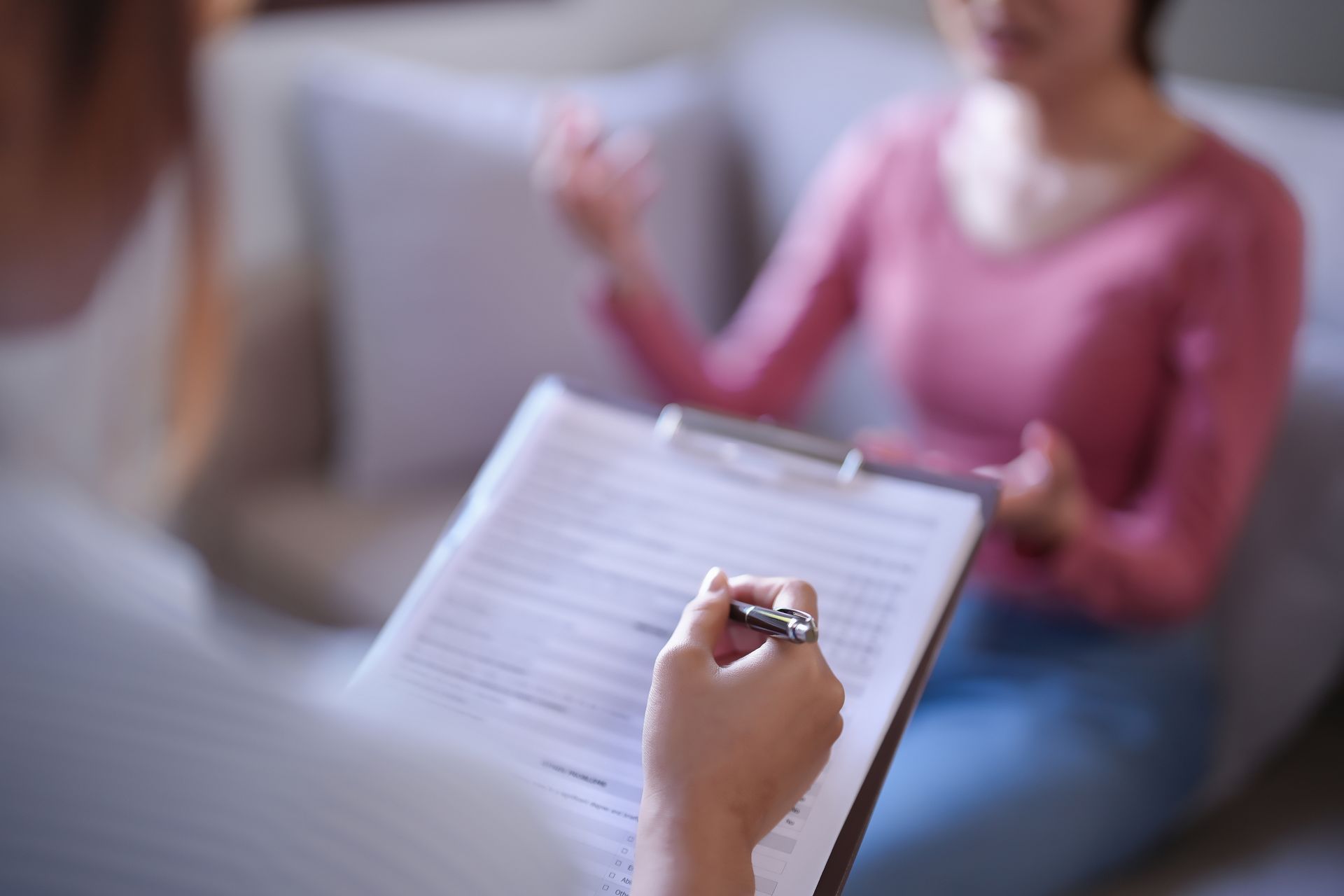 Hand writing on a clipboard while a blurred person sits across a table in the background