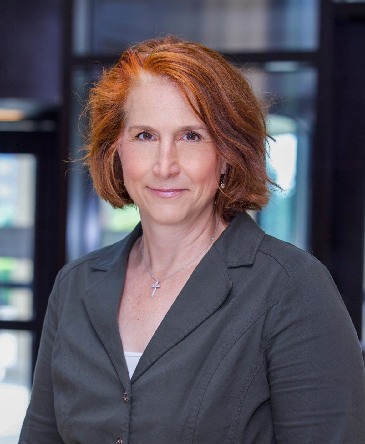 A woman with red hair is wearing a grey jacket and a cross necklace.
