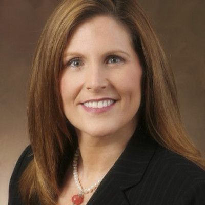 A woman wearing a black jacket and a red heart necklace smiles for the camera.