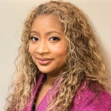 A woman with curly blonde hair is wearing a pink jacket and smiling for the camera.