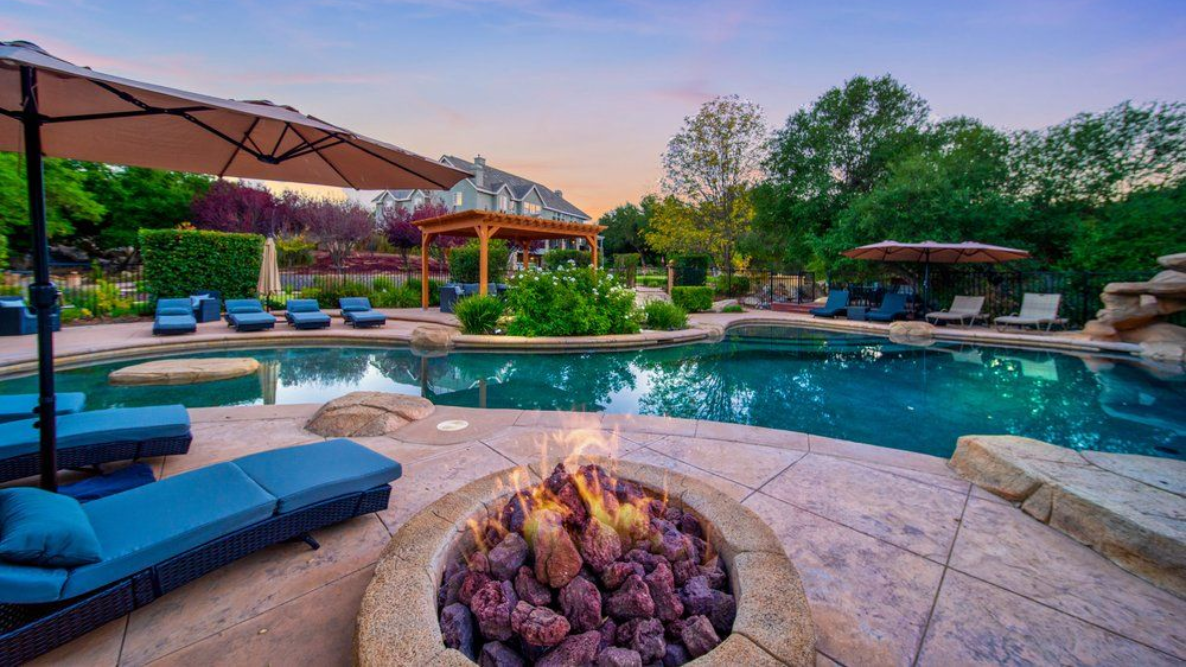 Luxury resort pool with fire pit at sunset, blue lounge chairs, umbrellas, and lush greenery