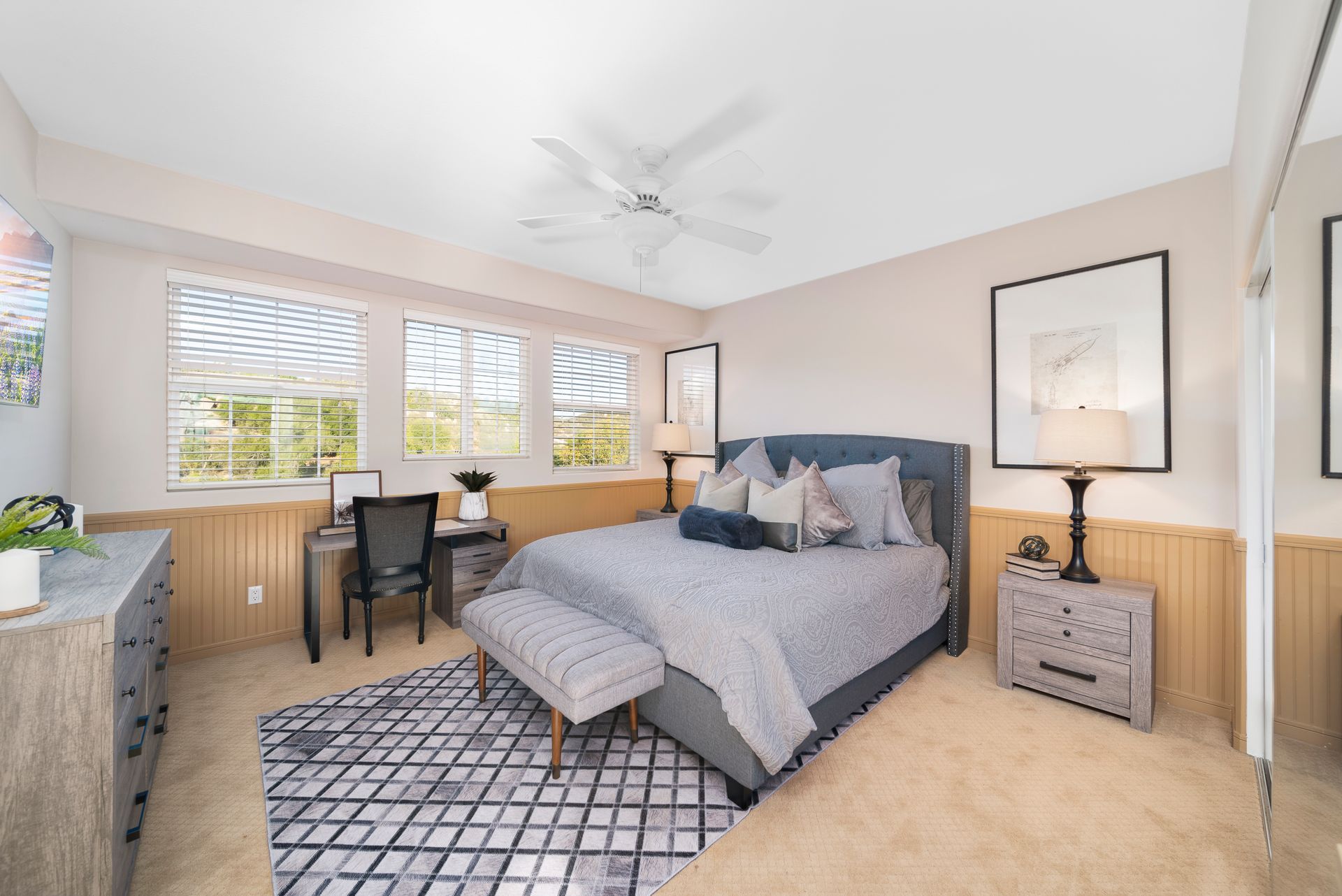 Bright bedroom with gray bed, desk, patterned bench, and large windows inviting daylight