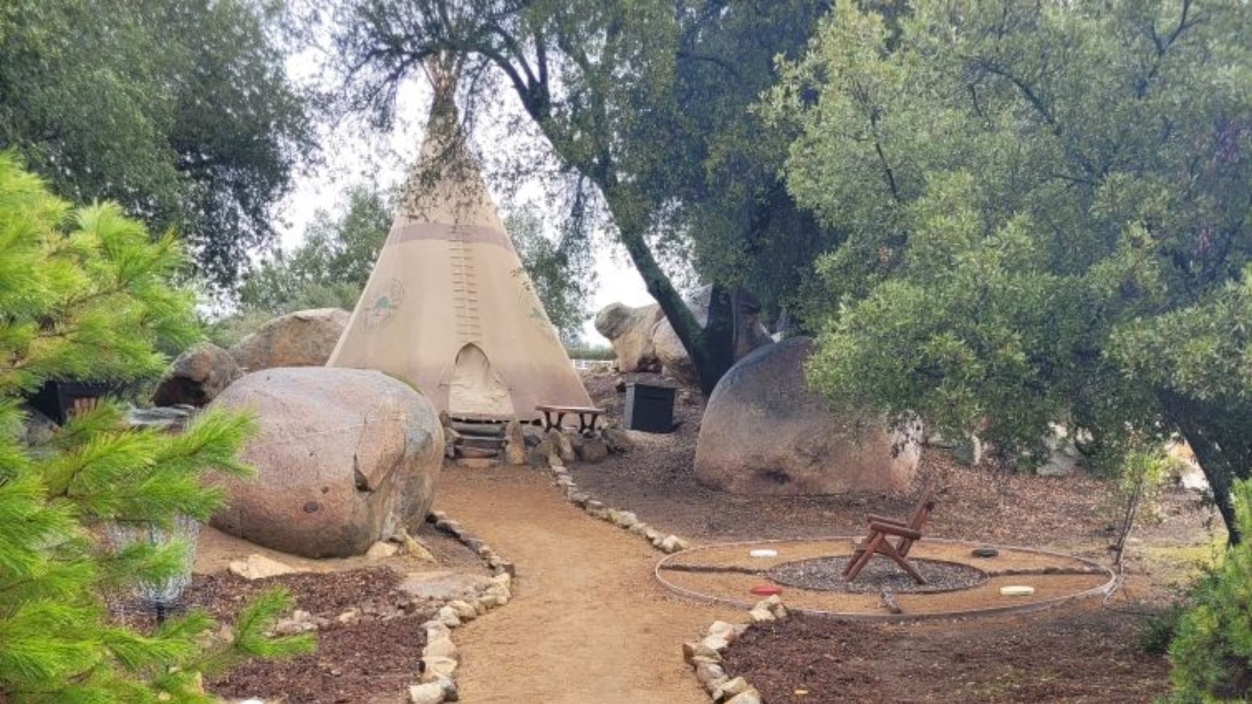 Outdoor tepee-style shelter among large rocks and trees on a dirt path in a wooded area