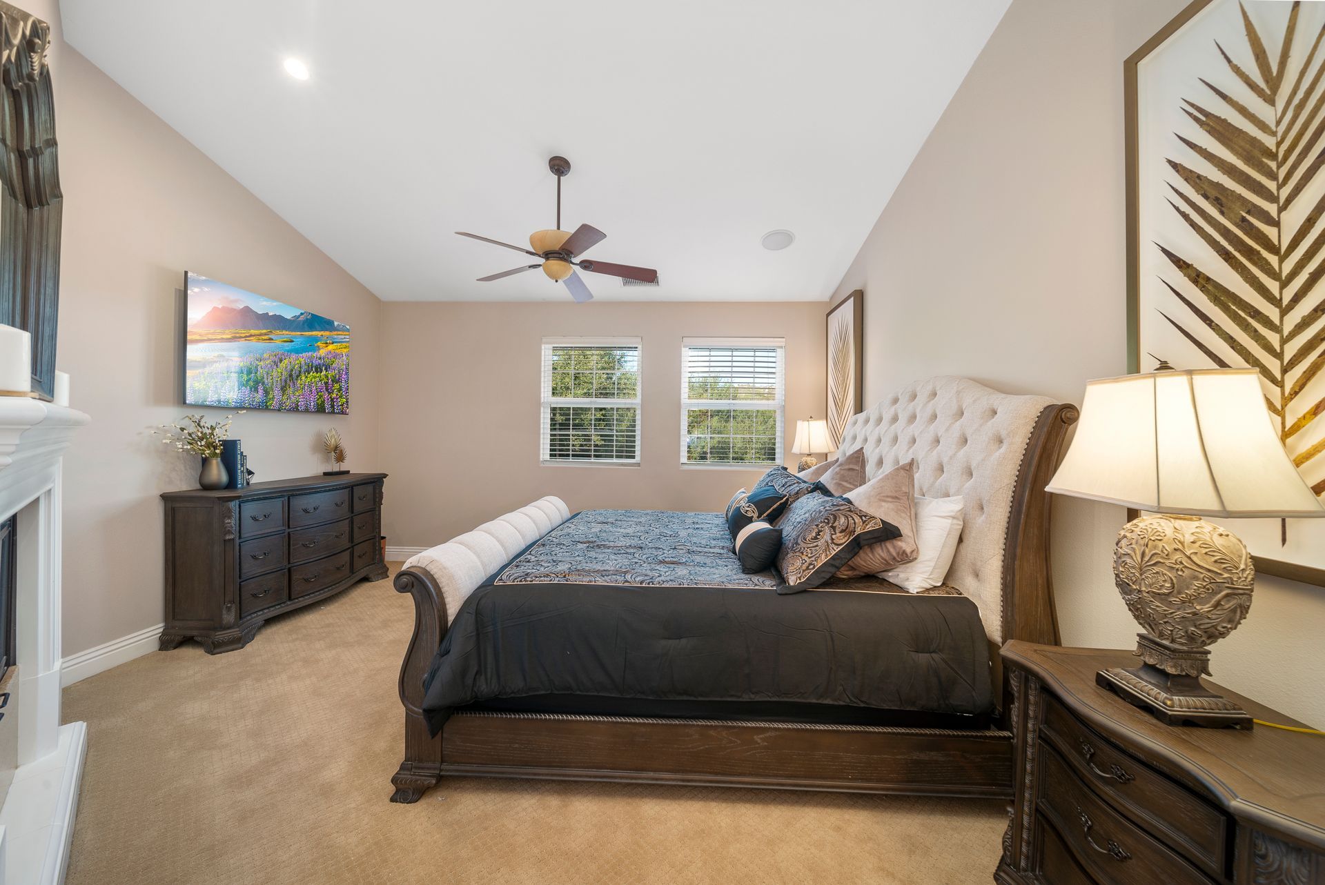 Bright bedroom with a tufted bed, dark dresser, nightstands, and a ceiling fan by two windows.