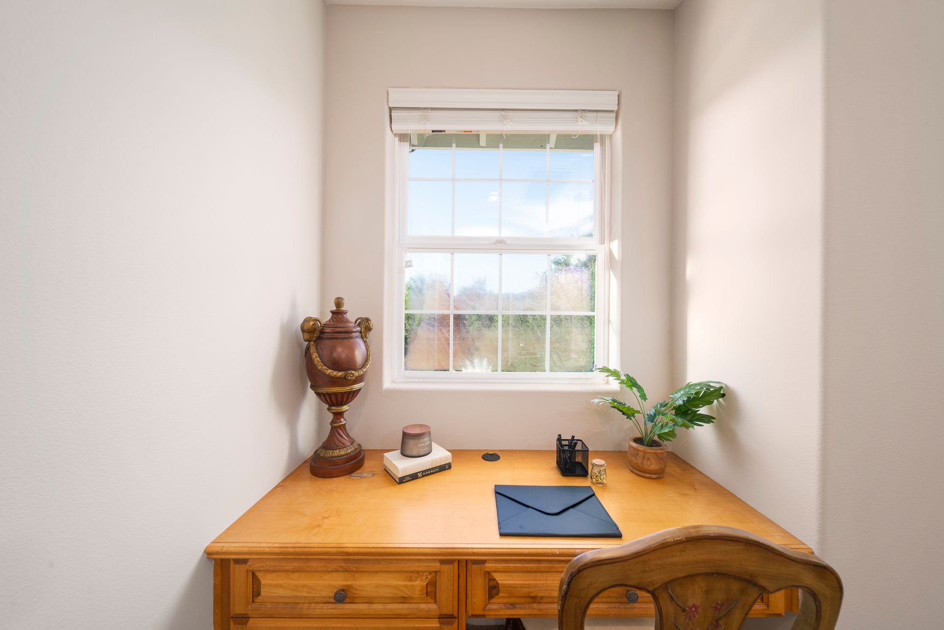 Small desk in a bright alcove under a window, with a vase, mug, notebook, and laptop on top.