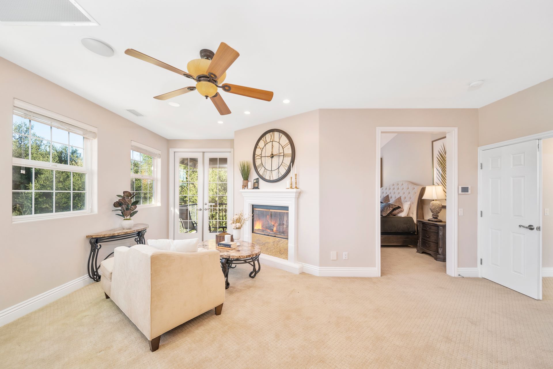 Bright living room with beige sofa, fireplace, ceiling fan, large windows, and open doorway to a bedroom.