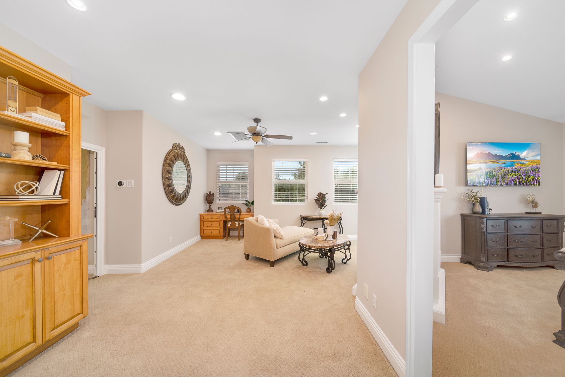 Bright living room with beige carpet, built-in wood shelves, and a small seating area by large windows.