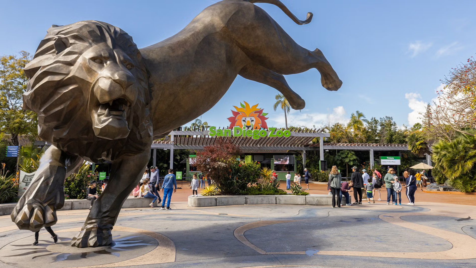Bronze lion sculpture rearing at a zoo entrance, with people walking in the sunny plaza.