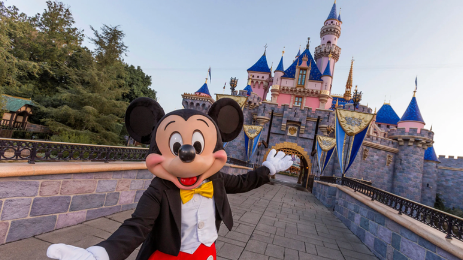 Mickey Mouse posing in front of a blue-and-gold castle at Disneyland, holding the camera for a selfie
