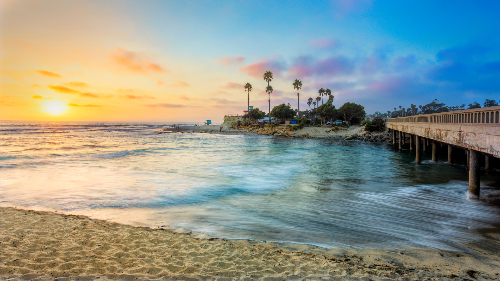 a sunset over a beach with a pier in the foreground and palm trees in the background .