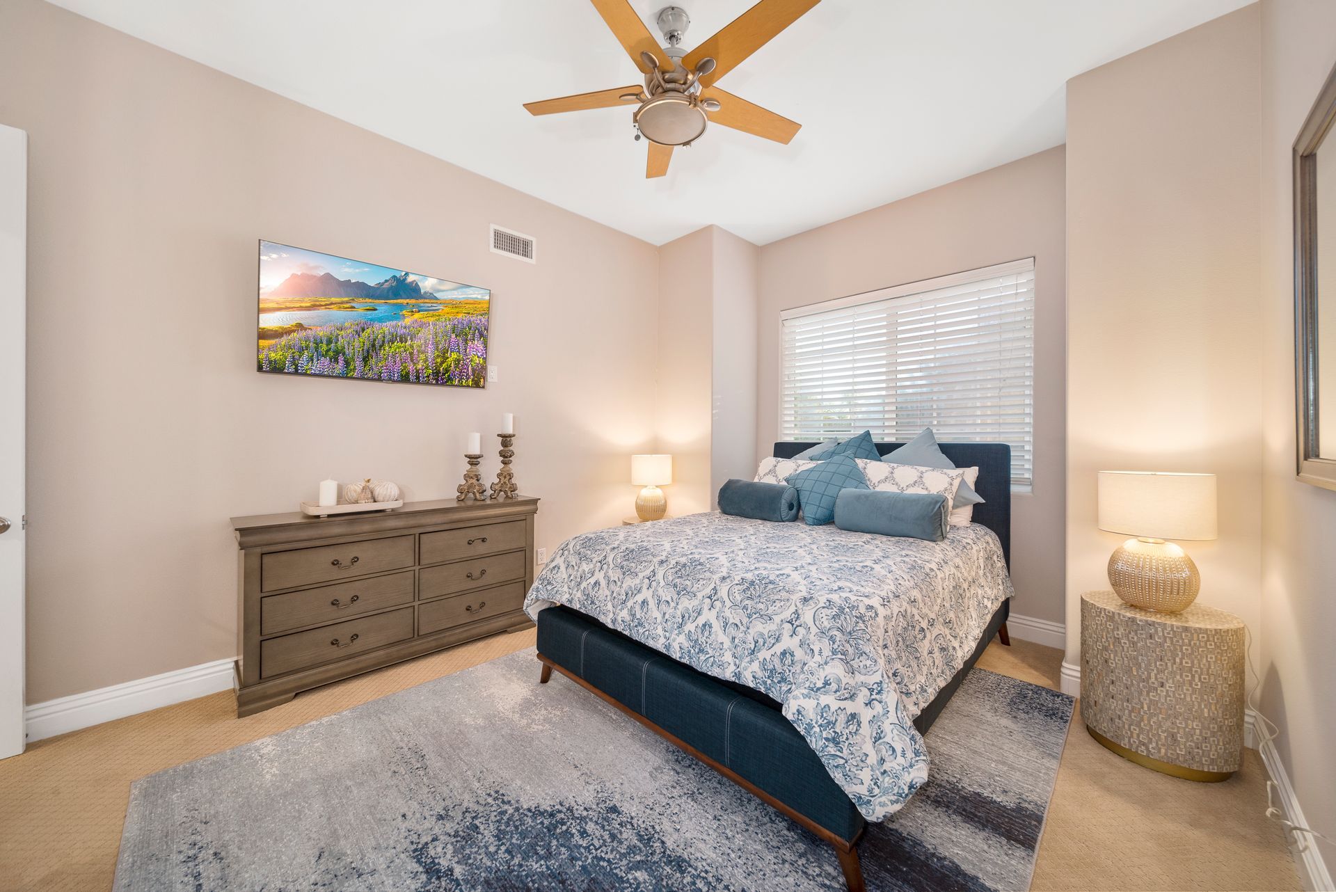 Bright bedroom with a bed, dresser, nightstands, lamp, ceiling fan, and window blinds