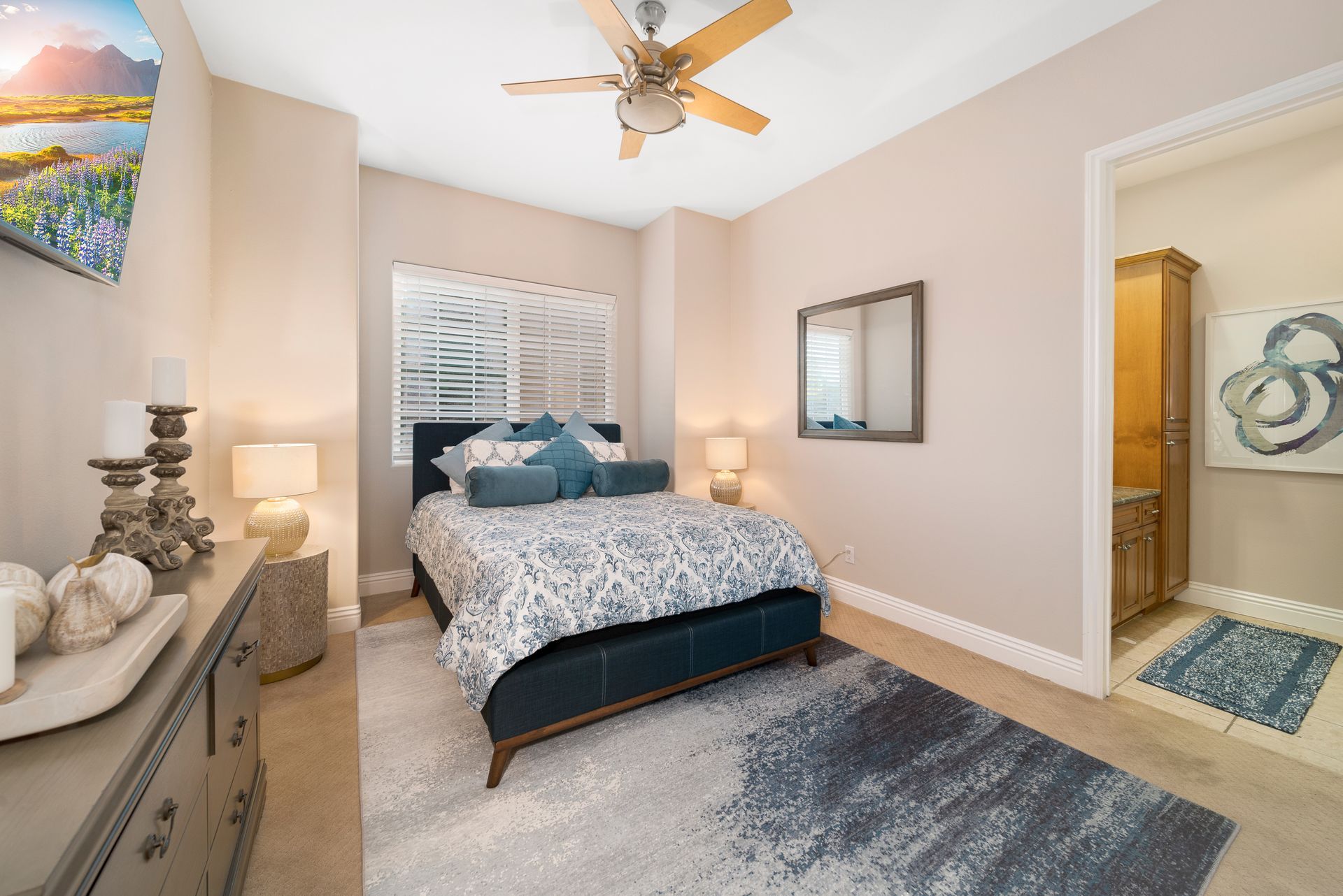 Bright bedroom with a bed, blue accents, ceiling fan, dresser, and bathroom doorway