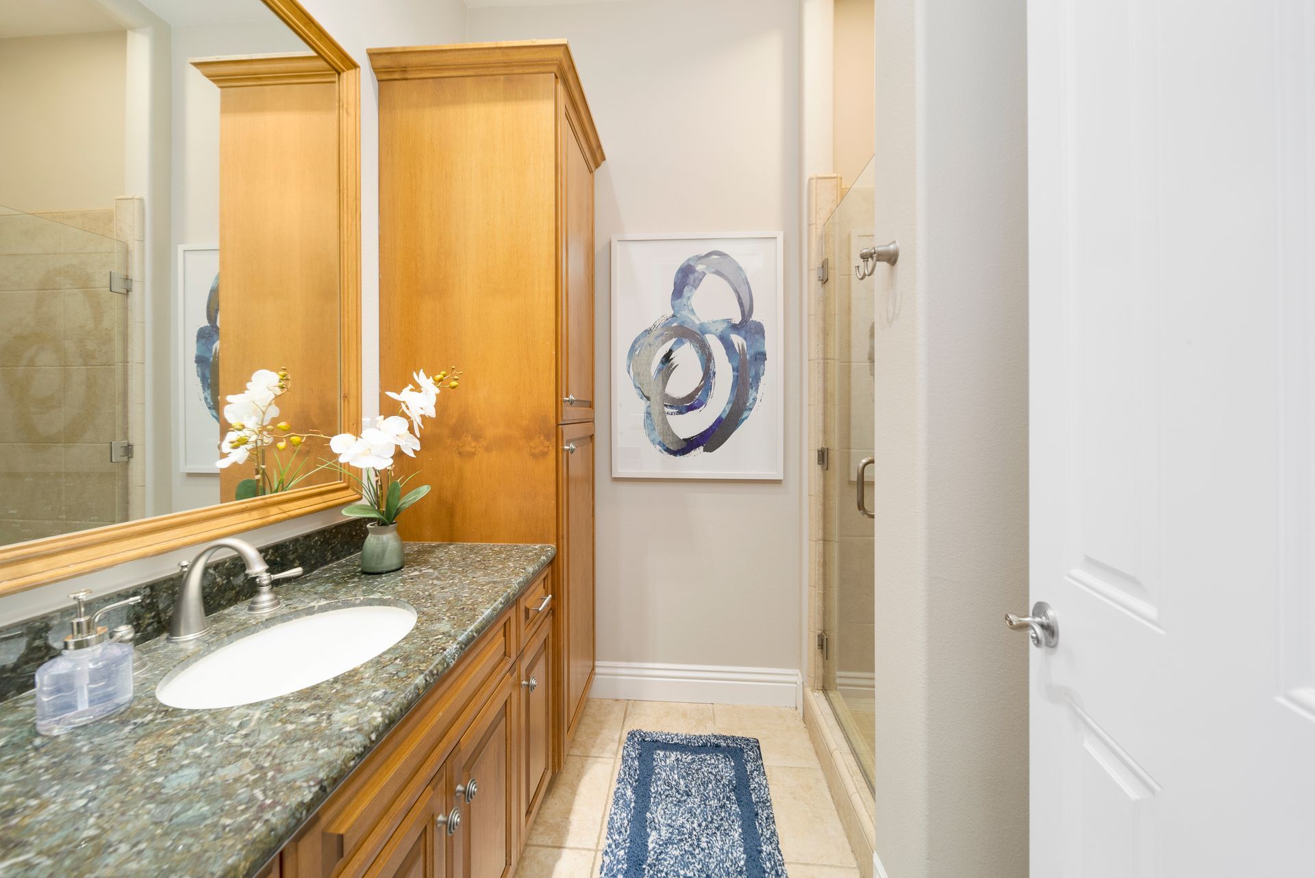 Bright bathroom with granite vanity, wooden cabinets, mirror, shower, and blue runner rug.