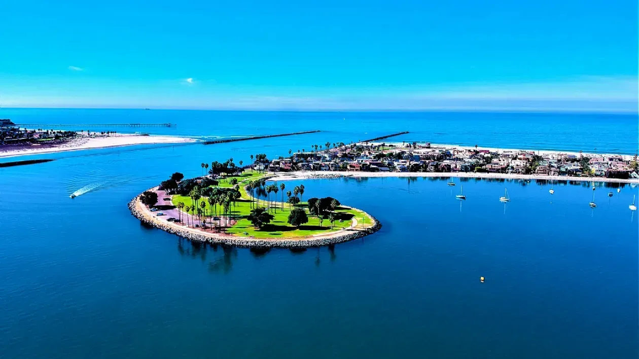 Aerial view of a green island in a calm blue bay with boats and a distant marina under clear sky