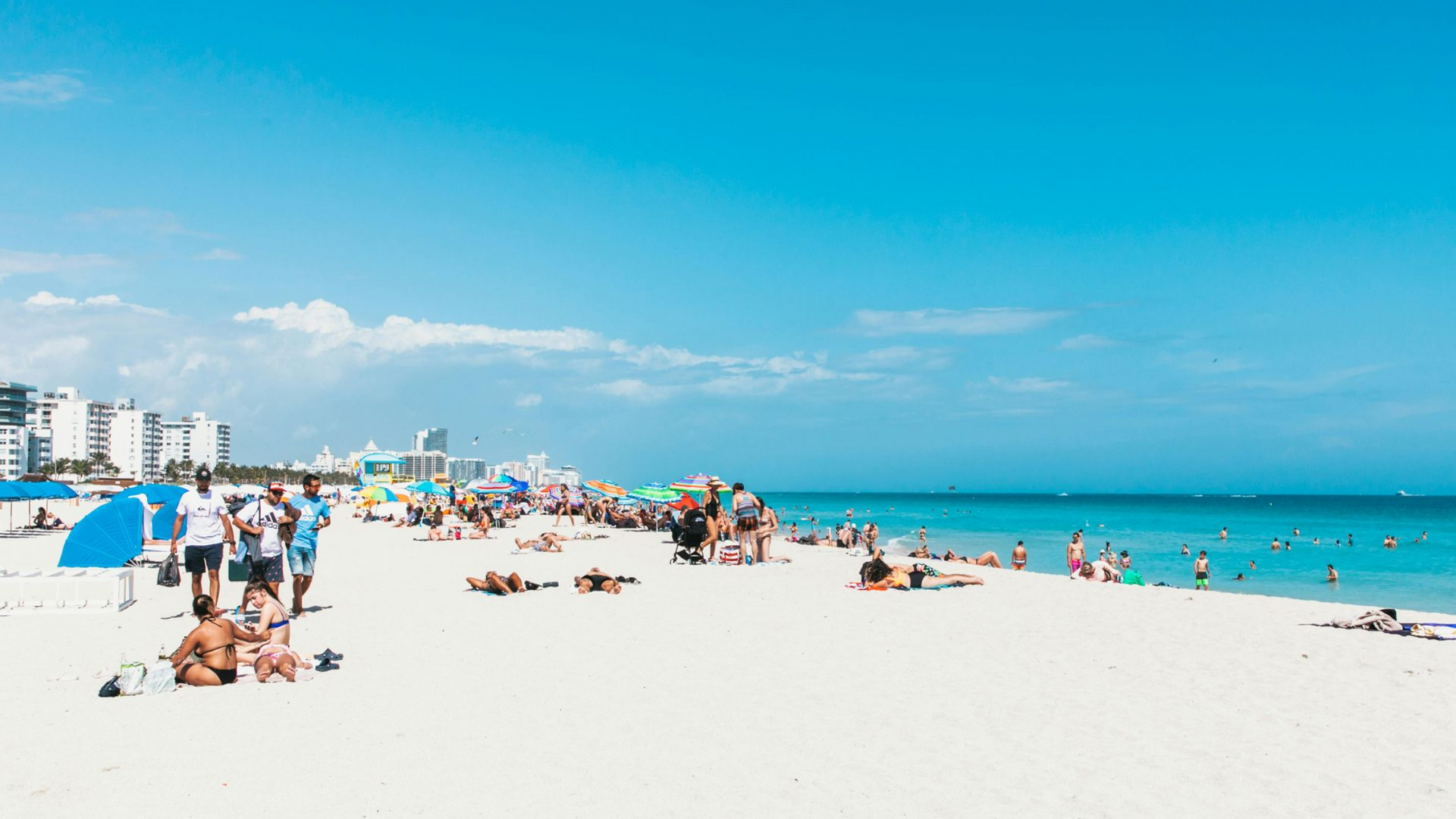 Sunny beach with people relaxing on white sand, turquoise water, and city buildings in the distance