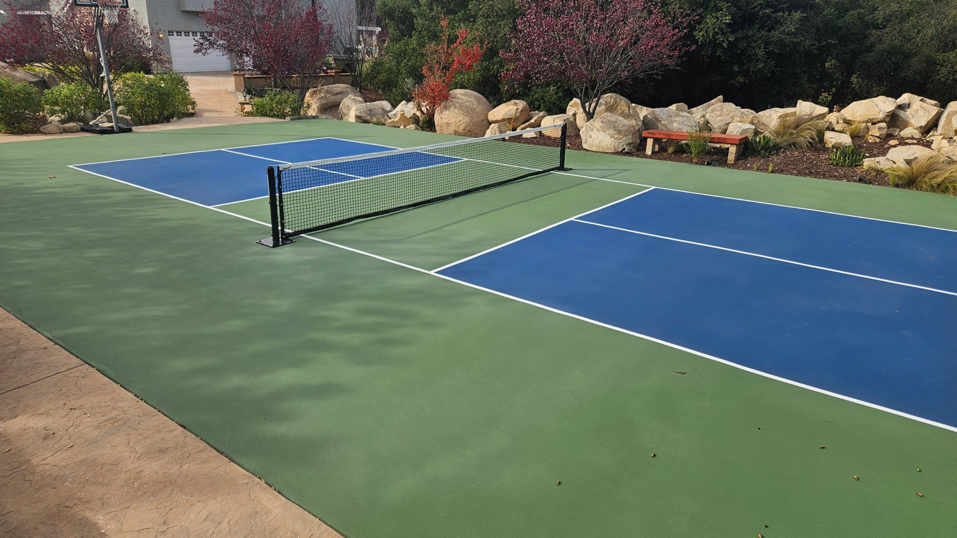 Outdoor pickleball court with blue playing surfaces, green surrounds, and a net between the two sides.