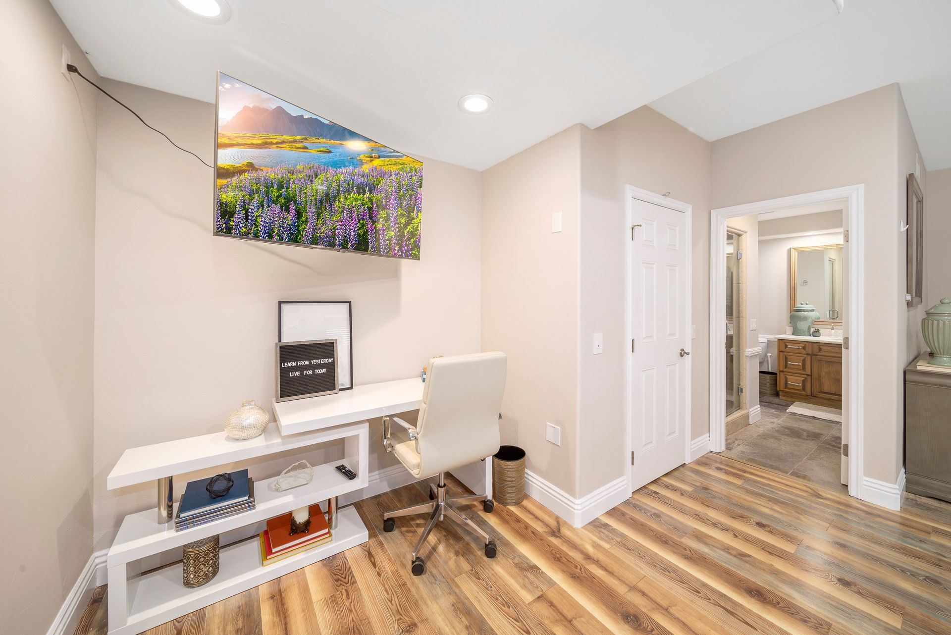 Home office with white desk and chair, colorful wall art, and wood flooring leading to a hallway with a bathroom.