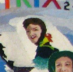 Smiling person in winter gear with a snowboard, near another person in the corner