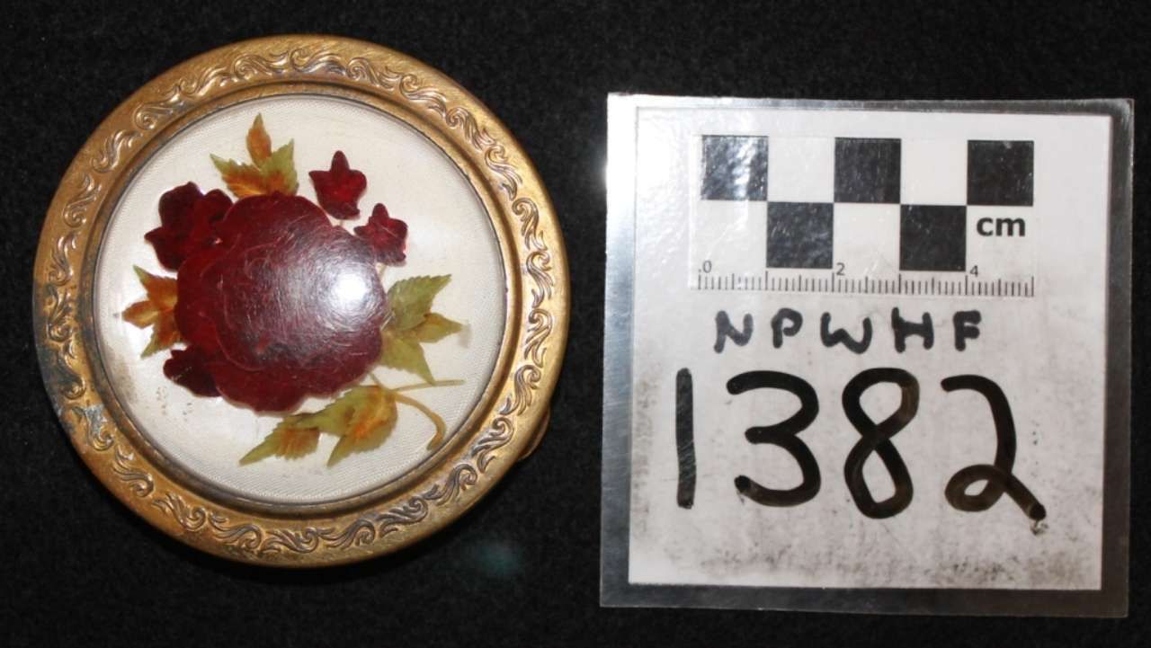 Round decorative brooch with red center and leaf motif beside a labeled scale card marked 1382