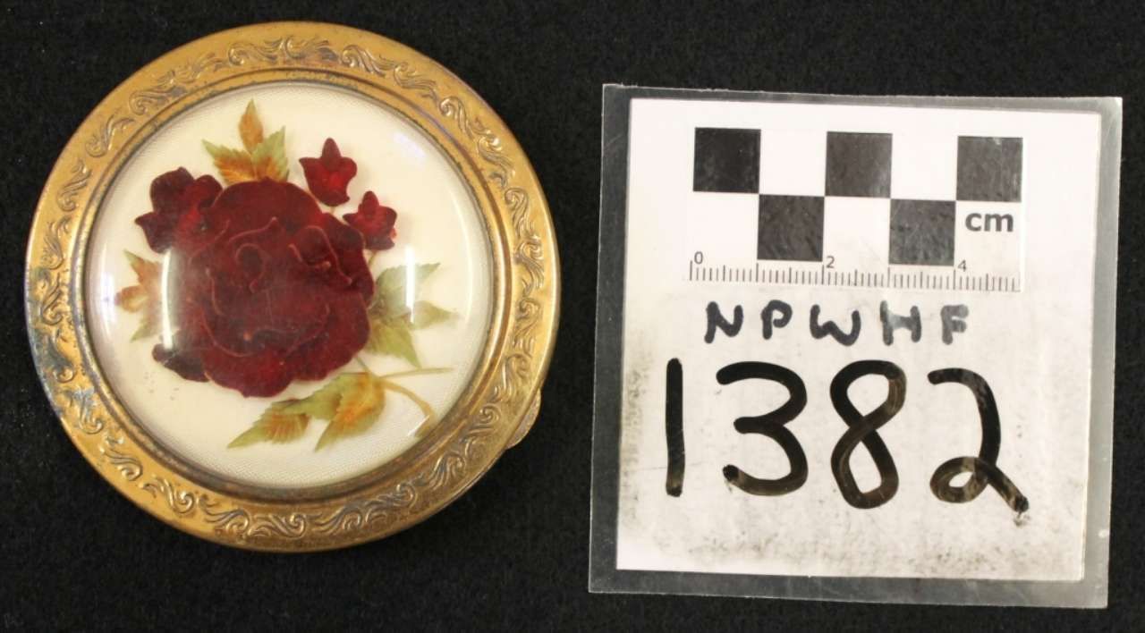 Round gold pin with red rose motif beside a labeled measurement card reading 1382