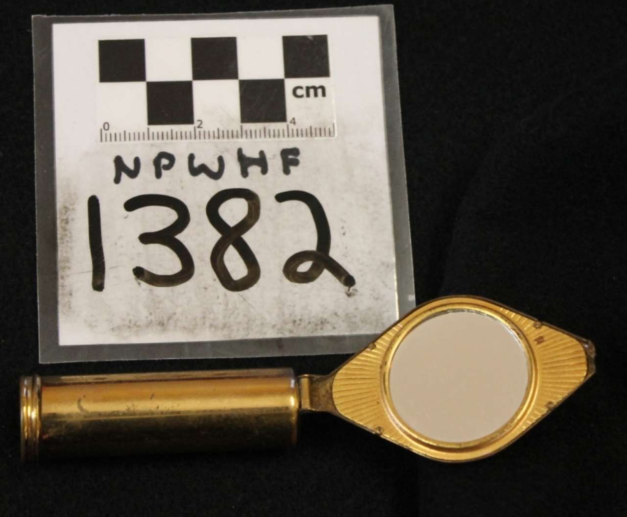 Brass-colored handheld magnifier or flashlight beside a labeled evidence scale card on a dark background