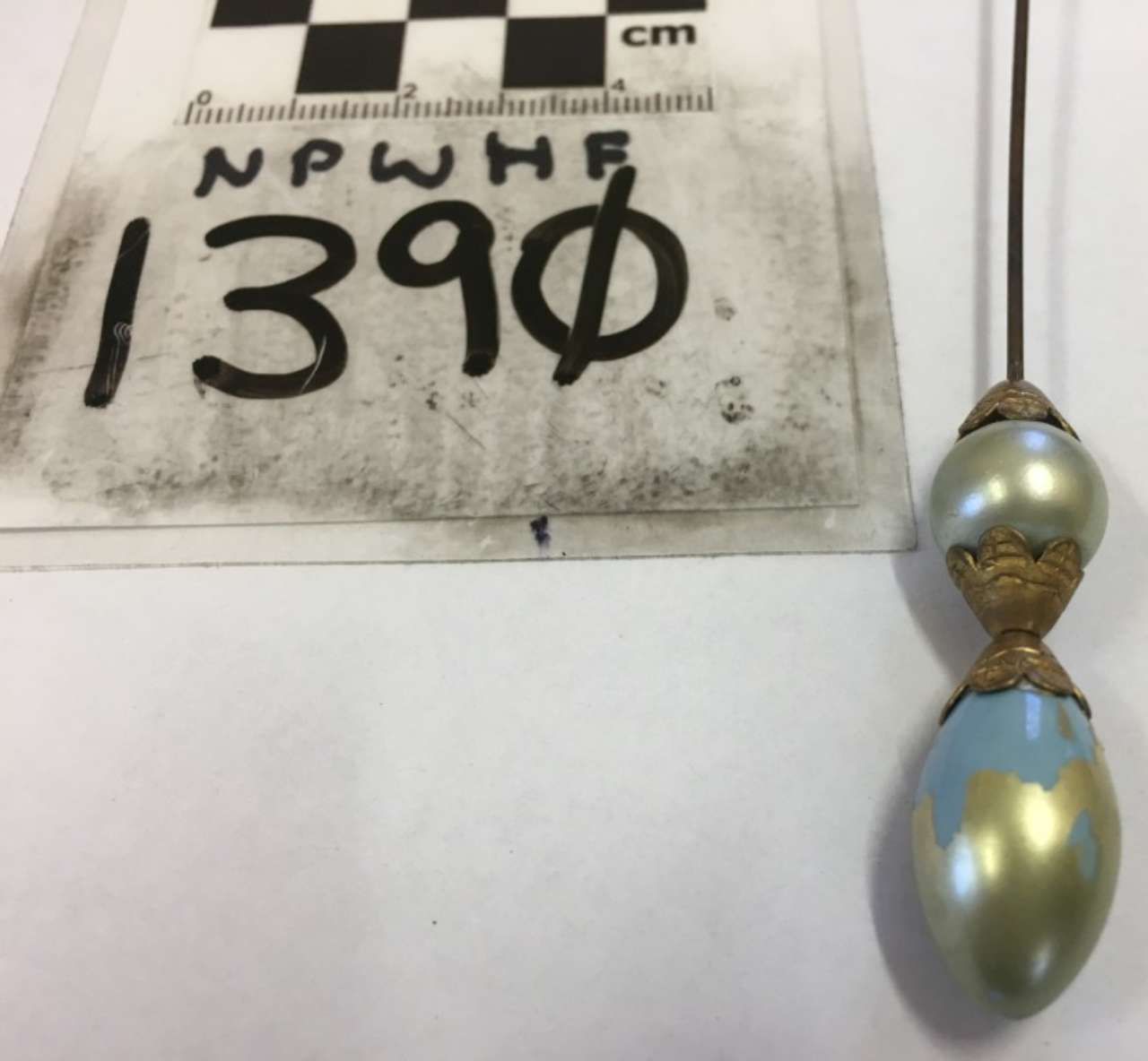 Small metallic teardrop pendant with gold band beside a labeled measurement sheet.