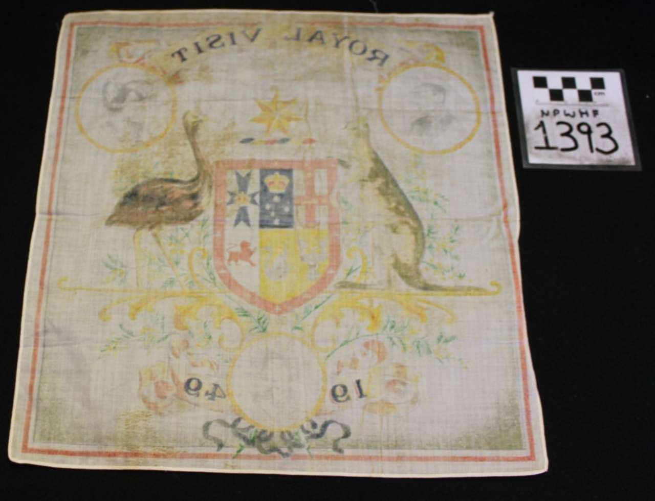 Faded embroidered crest on a pale cloth, with gold trim and a museum number tag beside it.