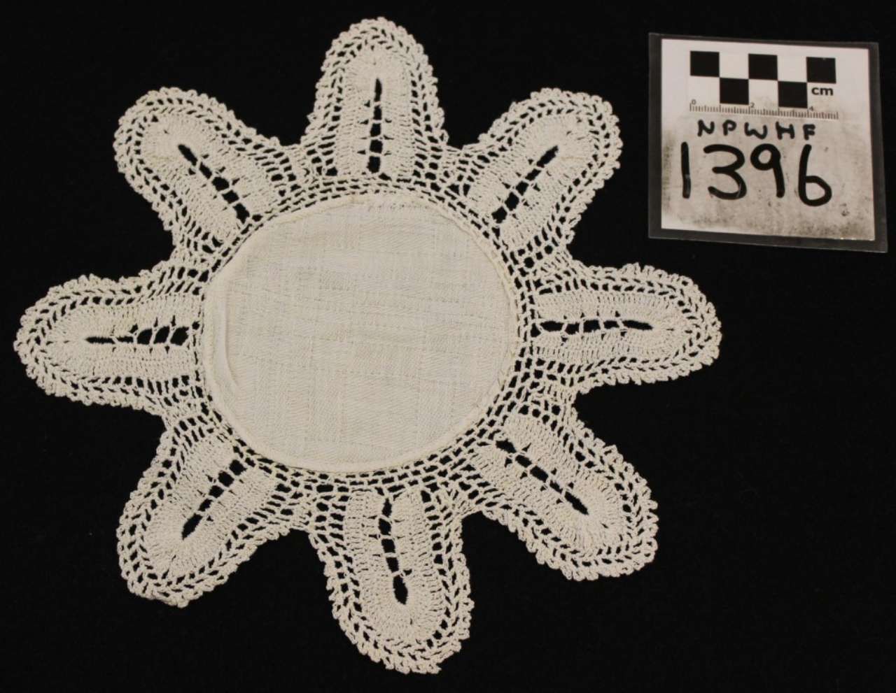White crocheted flower-shaped doily with eight petals beside a black-and-white label marked 1396.
