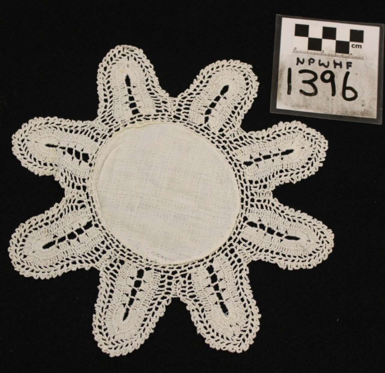 White crocheted lace doily with scalloped petals on a black background, beside a small numbered tag.
