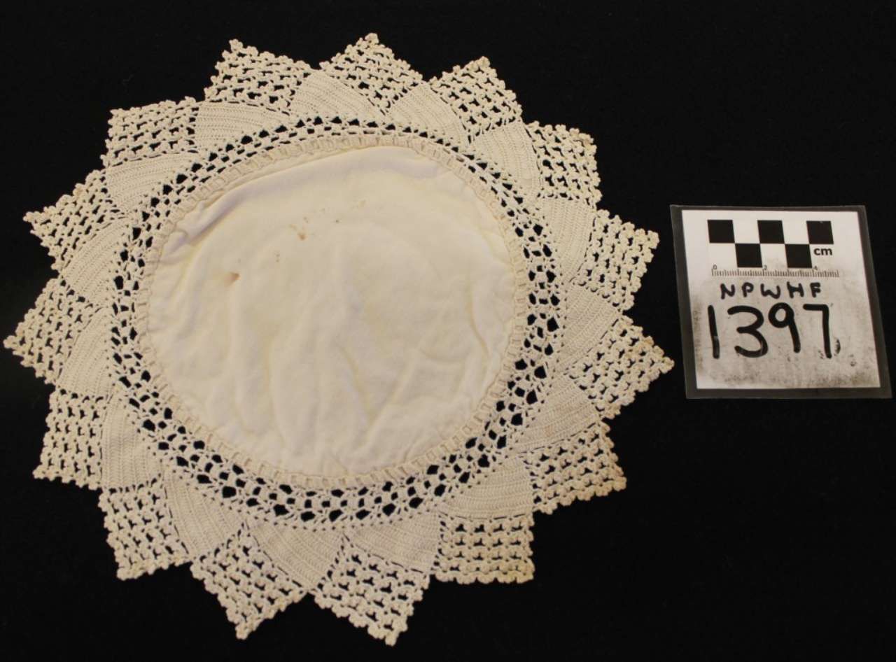 Beige crocheted lace doily with scalloped edges, displayed beside a numbered museum tag.