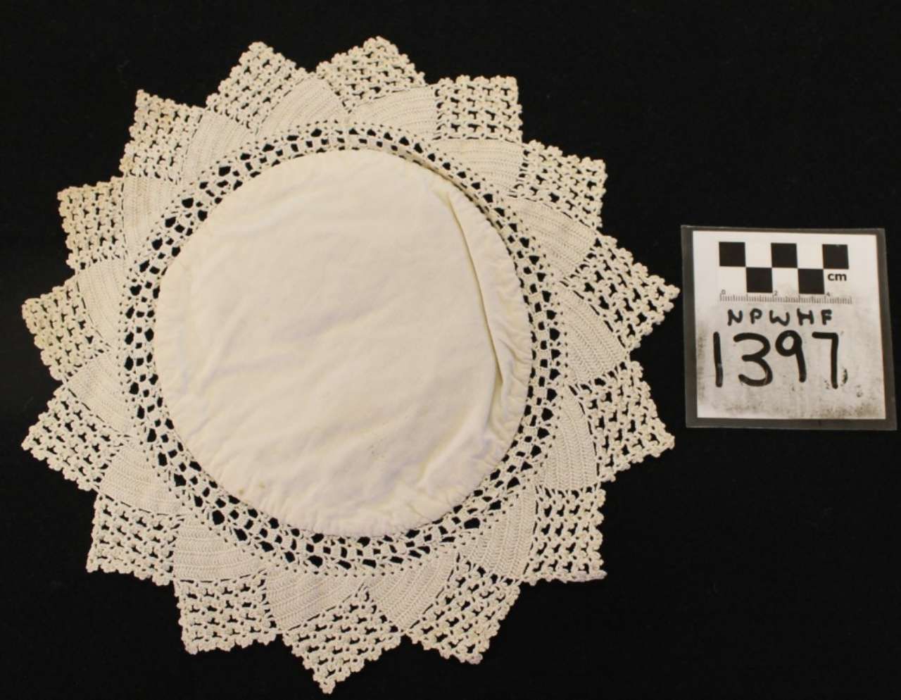 White crocheted lace doily with scalloped edges on black background, beside a small numbered label.
