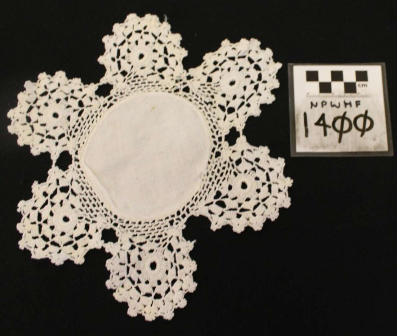 White lace doily with six floral points on black background, beside a small labeled scale card.