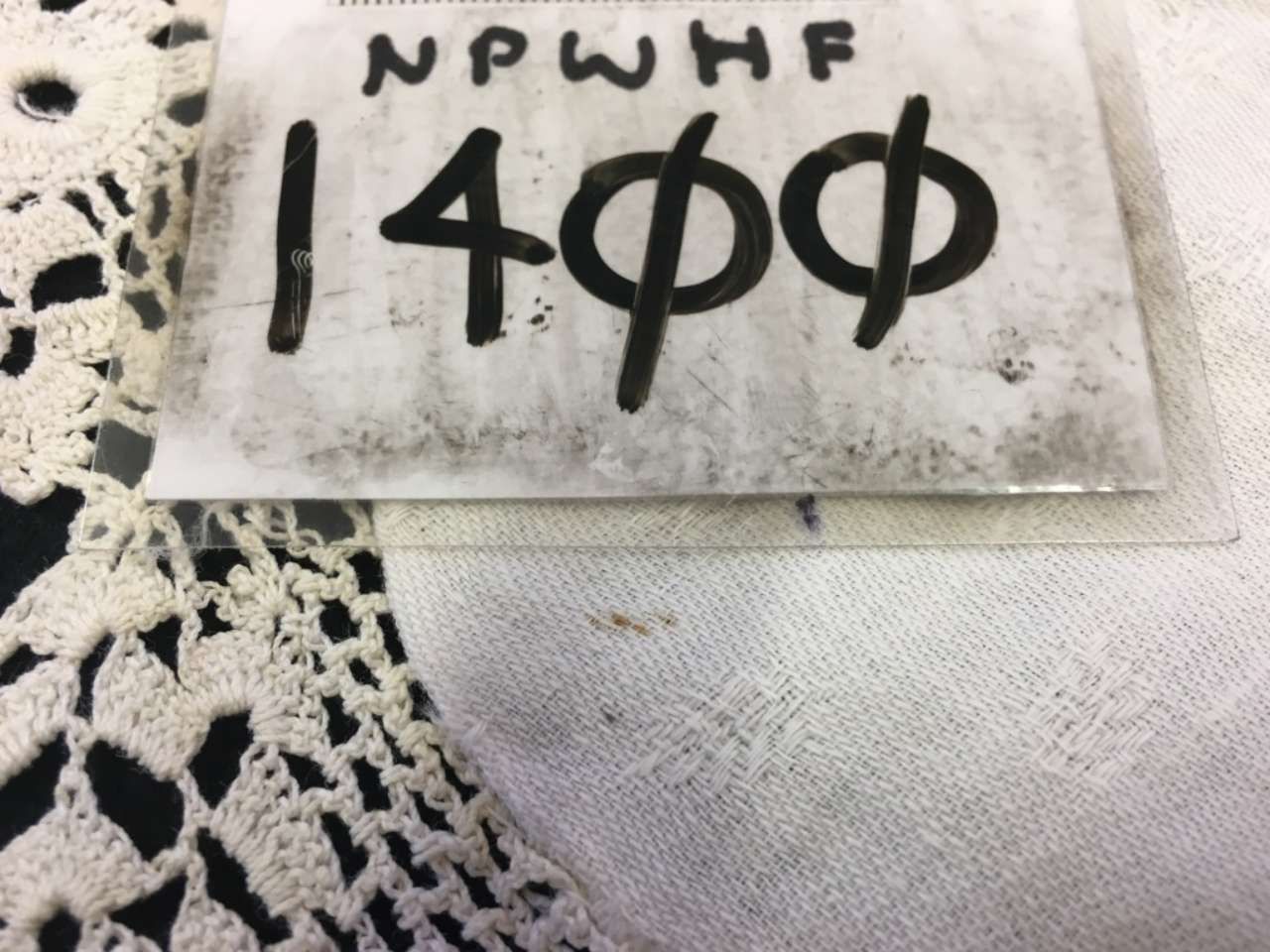 White fabric tag with black handwritten text “NPWHF 1400,” beside a crocheted lace edge.