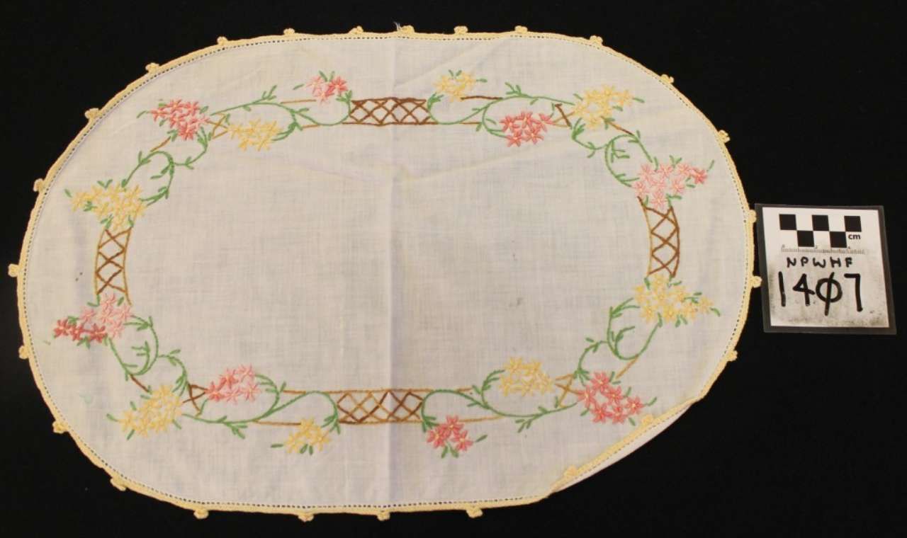 Oval embroidered tablecloth with floral border, shown beside a small label.