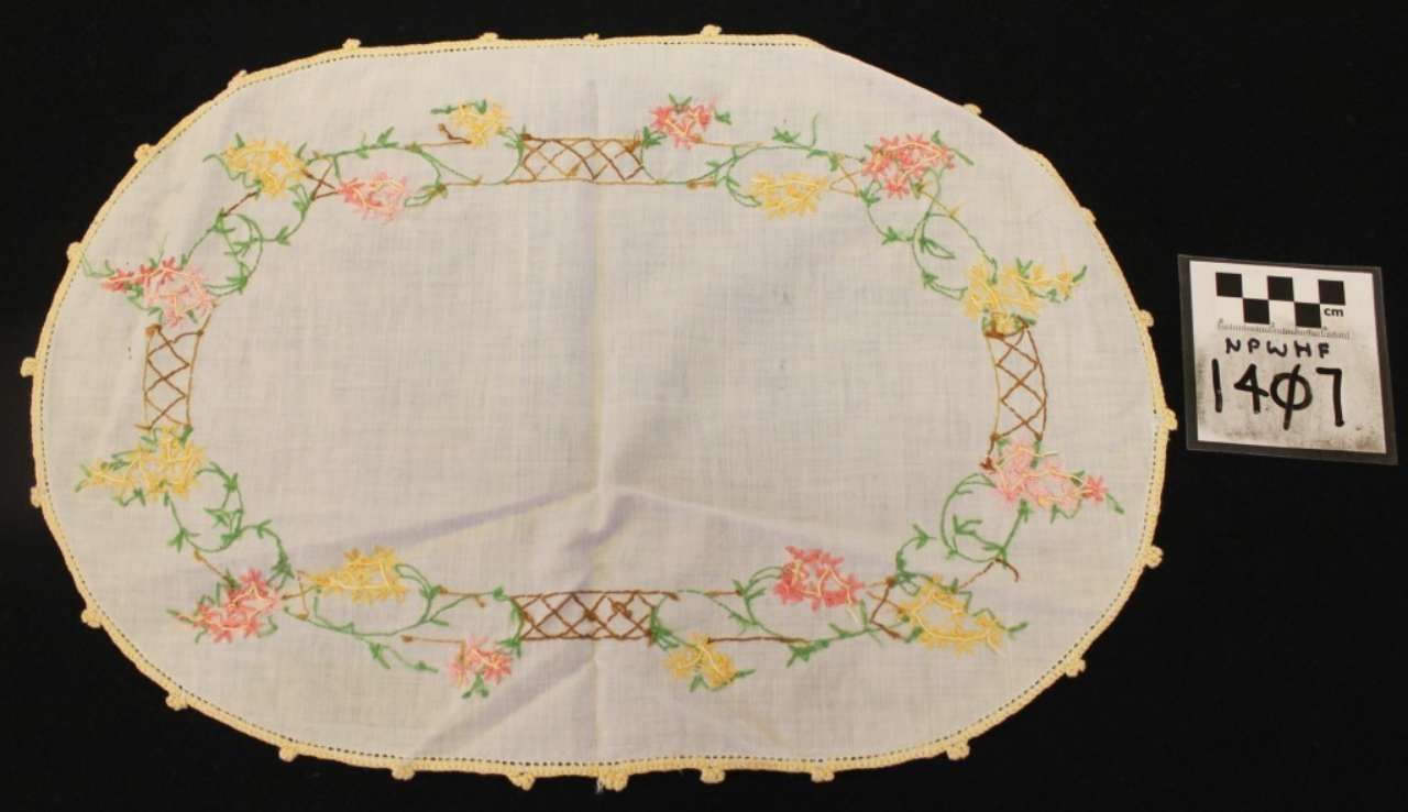Oval white embroidered doily with pastel floral wreath and scalloped border on black background