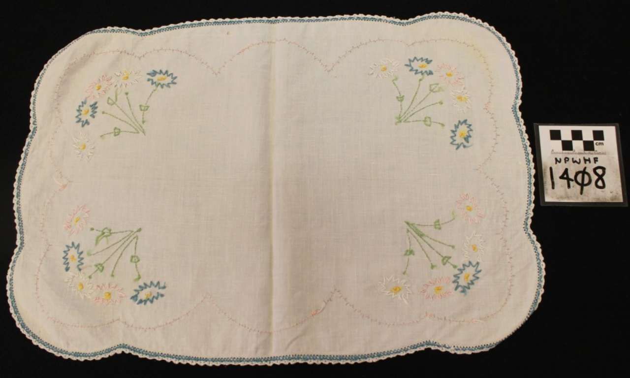 White embroidered placemat with scalloped edges and small pastel floral designs, shown on a black background