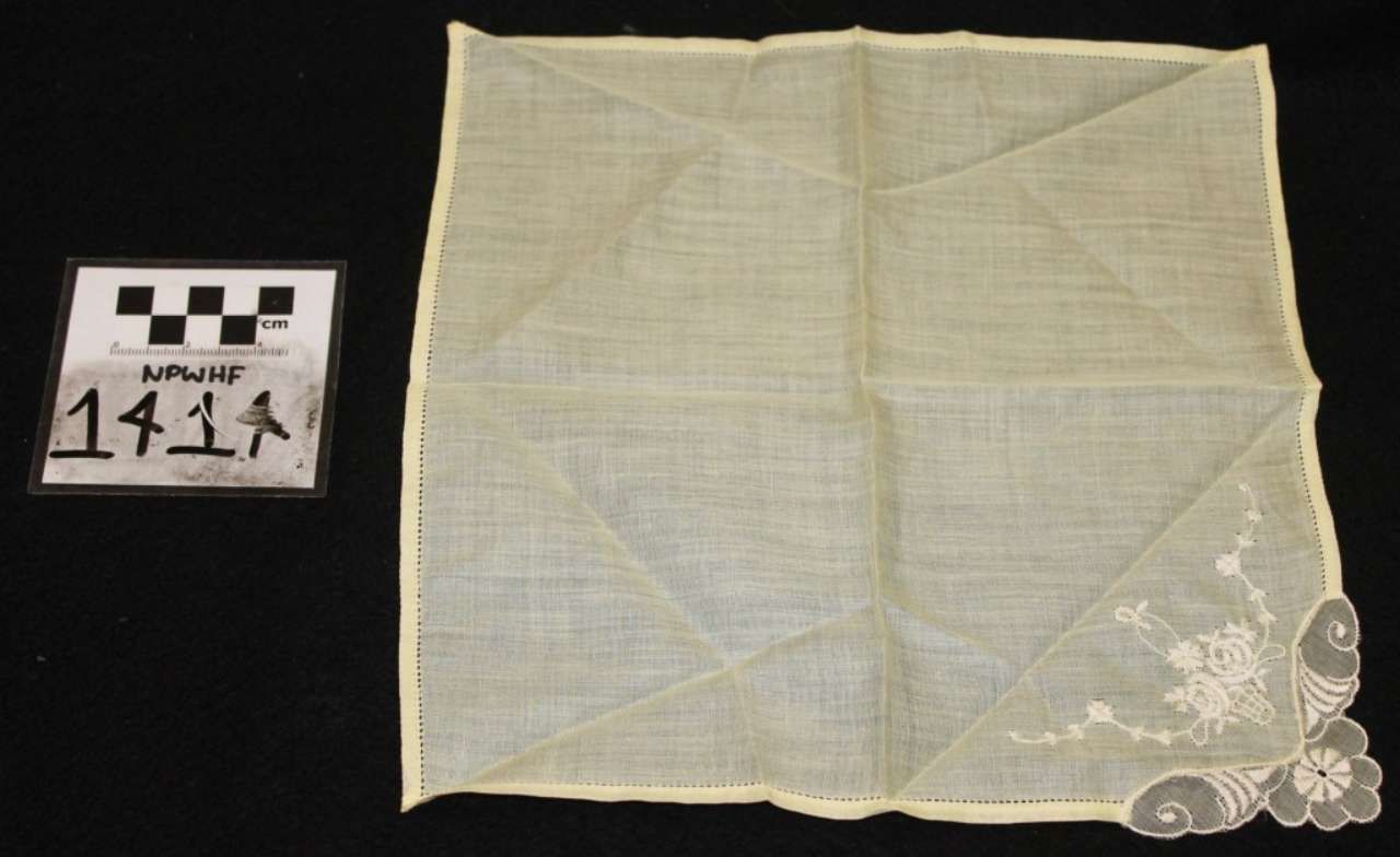 Cream lace-edged square handkerchief with a small black-and-white label beside it on a dark background