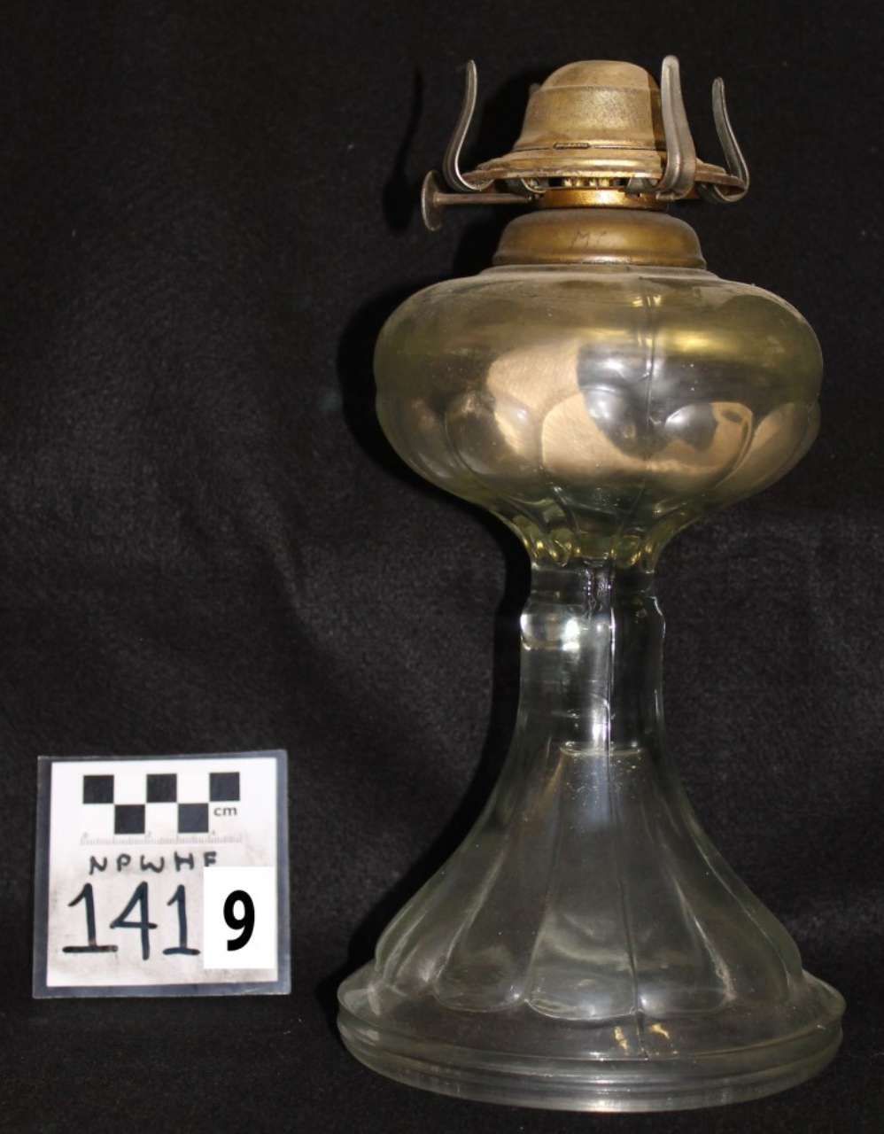 Vintage glass oil lamp with brass burner and clear chimney on a dark background, next to a museum ID card.