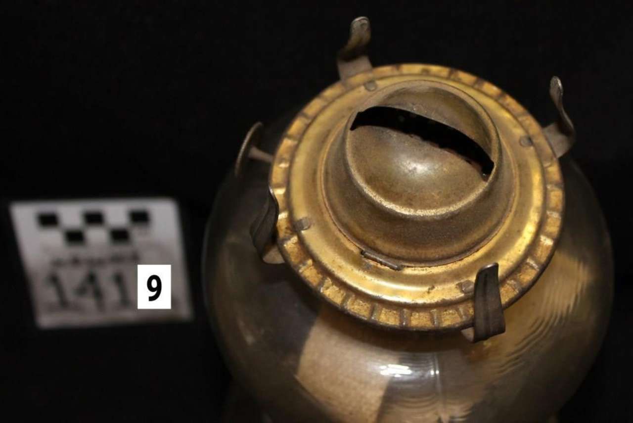 Close-up of a brass pot with a round lid and dark background, next to a small numbered tag.