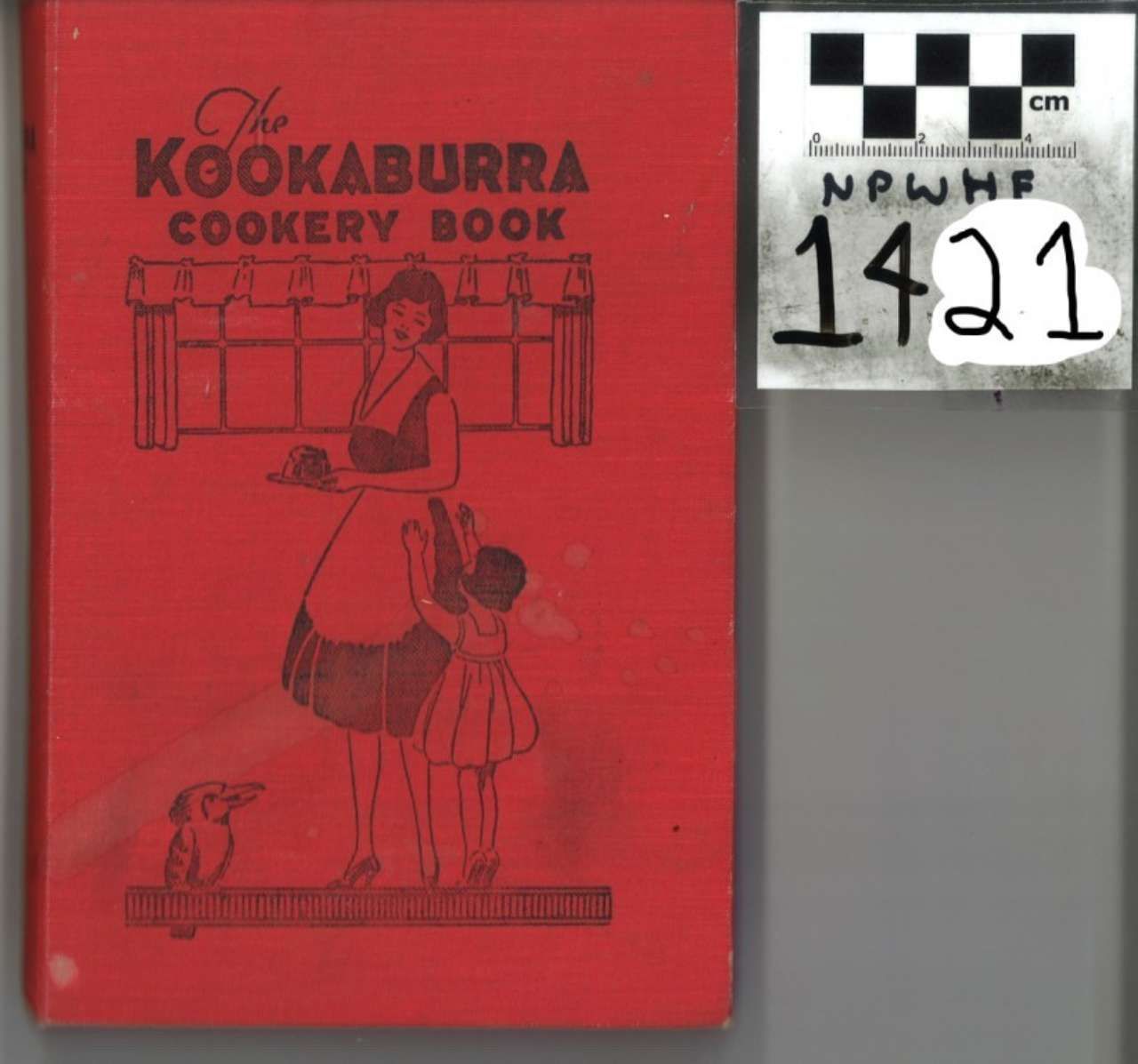 Red cookbook titled “The Kookaburra Cookery Book” beside a numbered archival label on gray background