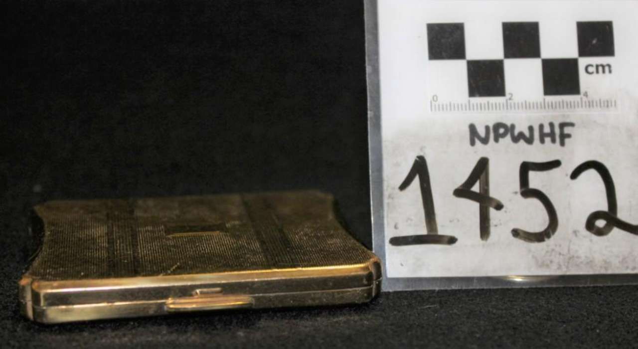 Small gold-toned metal lighter with etched top, beside a label marked “NPWHF 1452.”