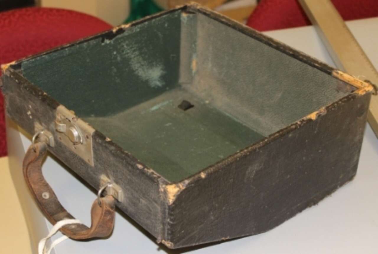 Small worn black metal box with a handle and green interior, sitting on a white surface.