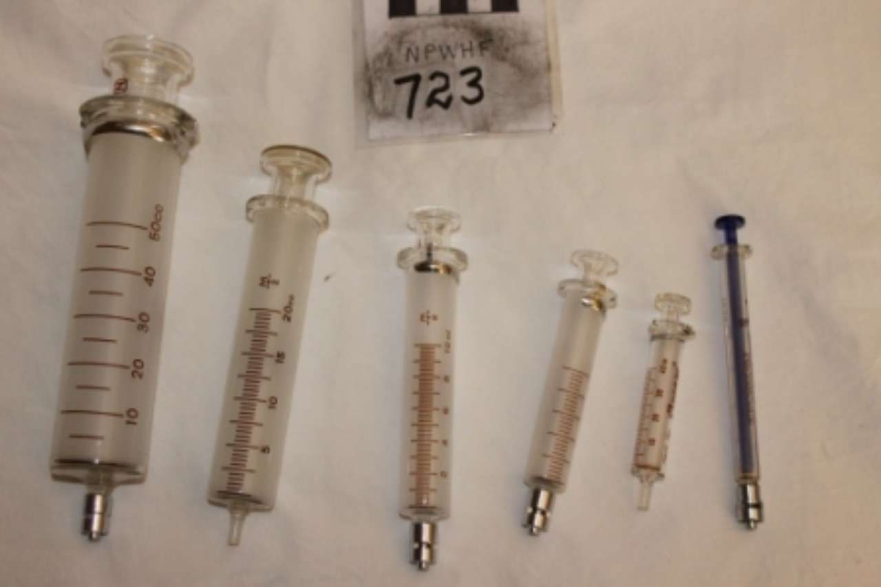 Several glass syringes of different sizes laid out on a white surface, with a small bag labeled 723 above them