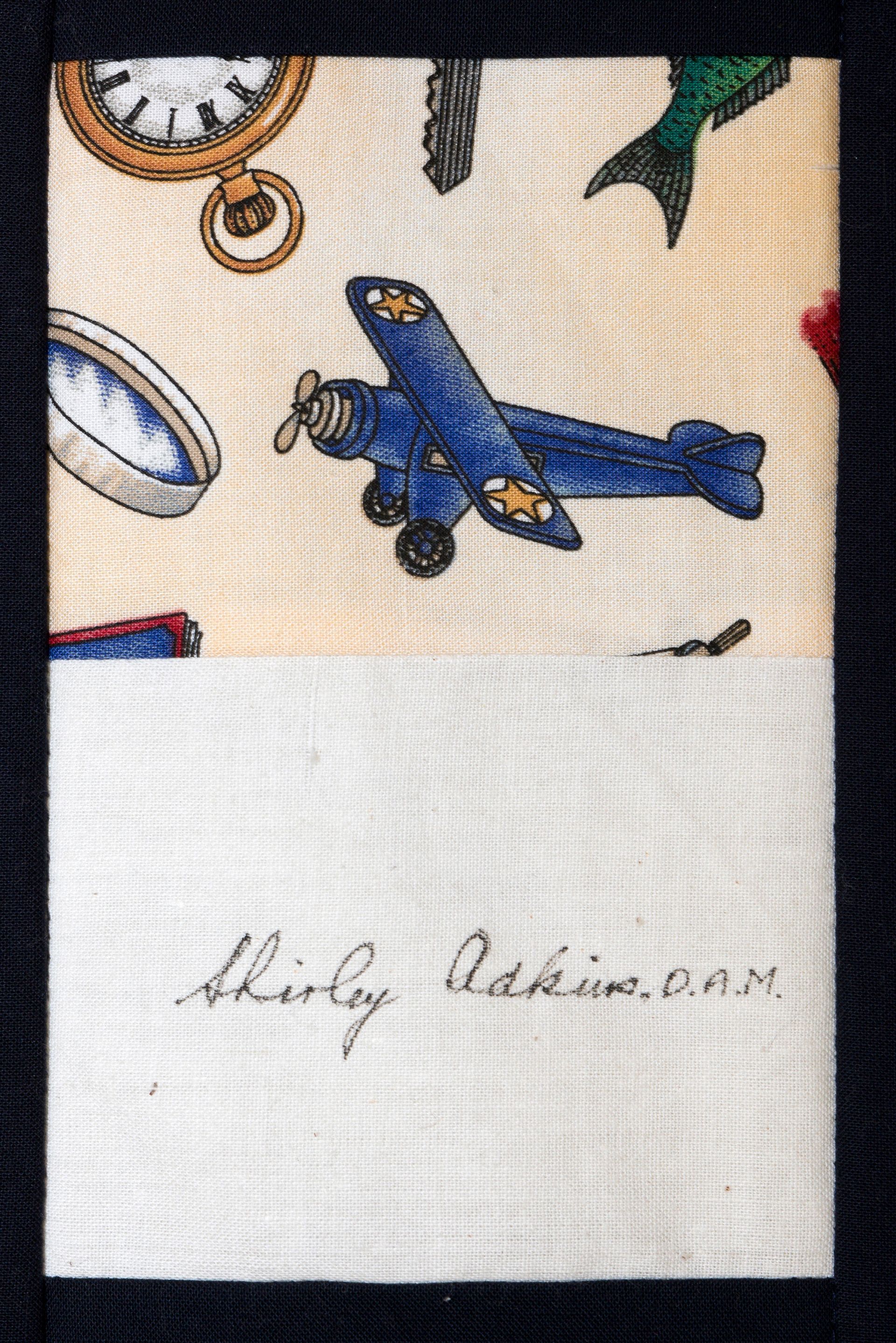 Blue toy airplane on patterned fabric above a white label with handwriting.